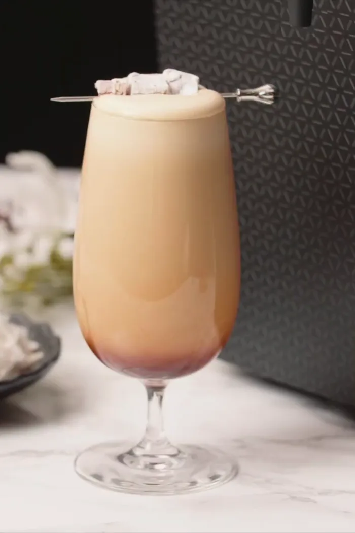 A Turkish delight cold brew in a tall glass with a toasted marshmallow skewer, beside a DRNX machine