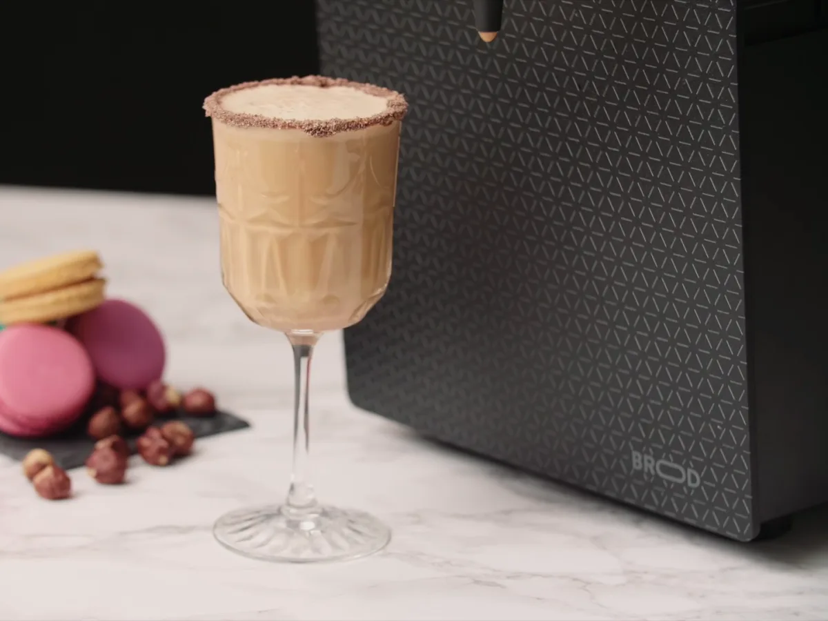 A hazelnut latte with a chocolate rim beside a plate of macarons and a DRNX machine